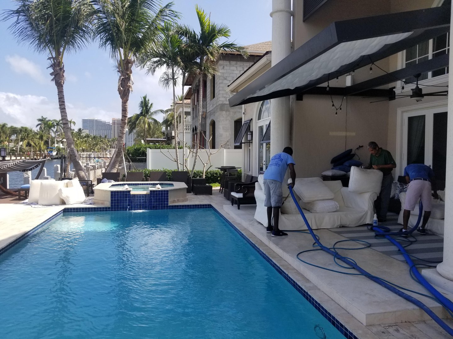 Dolphin team cleaning white outdoor sofas on the poolside patio of a waterfront home with palm trees