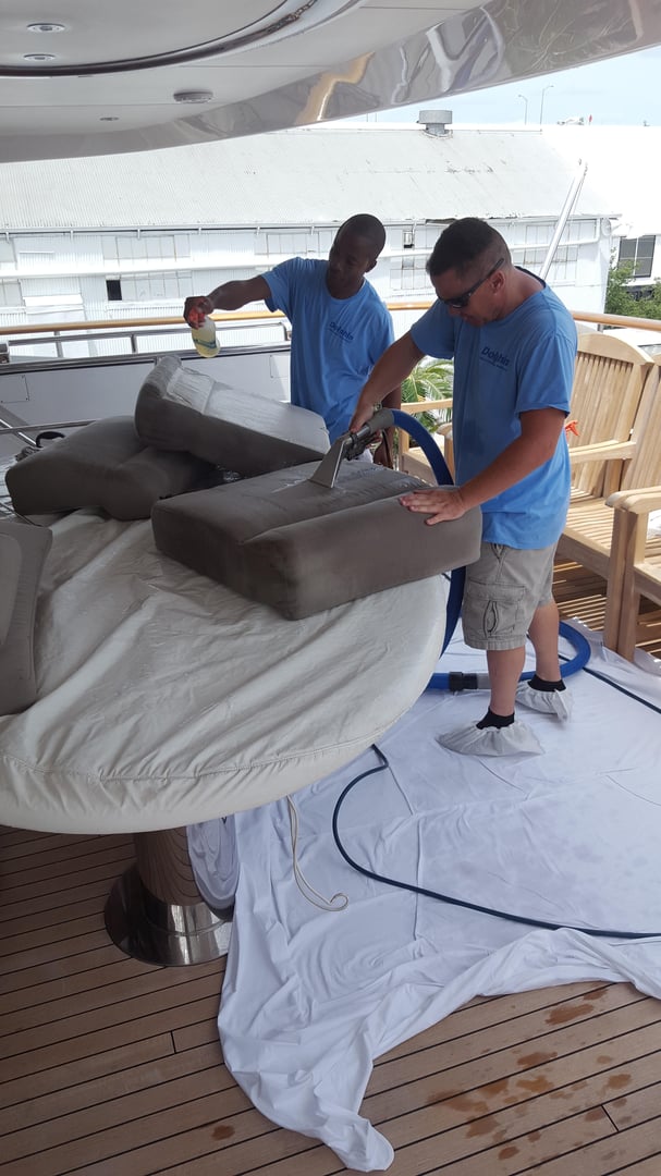 Two uniformed Dolphin technicians cleaning gray yacht deck cushions with an extraction wand on a protective sheet