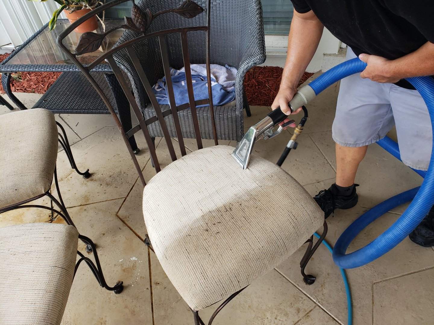 Technician cleaning a dirty upholstered patio chair seat with a handheld extraction tool and blue hose