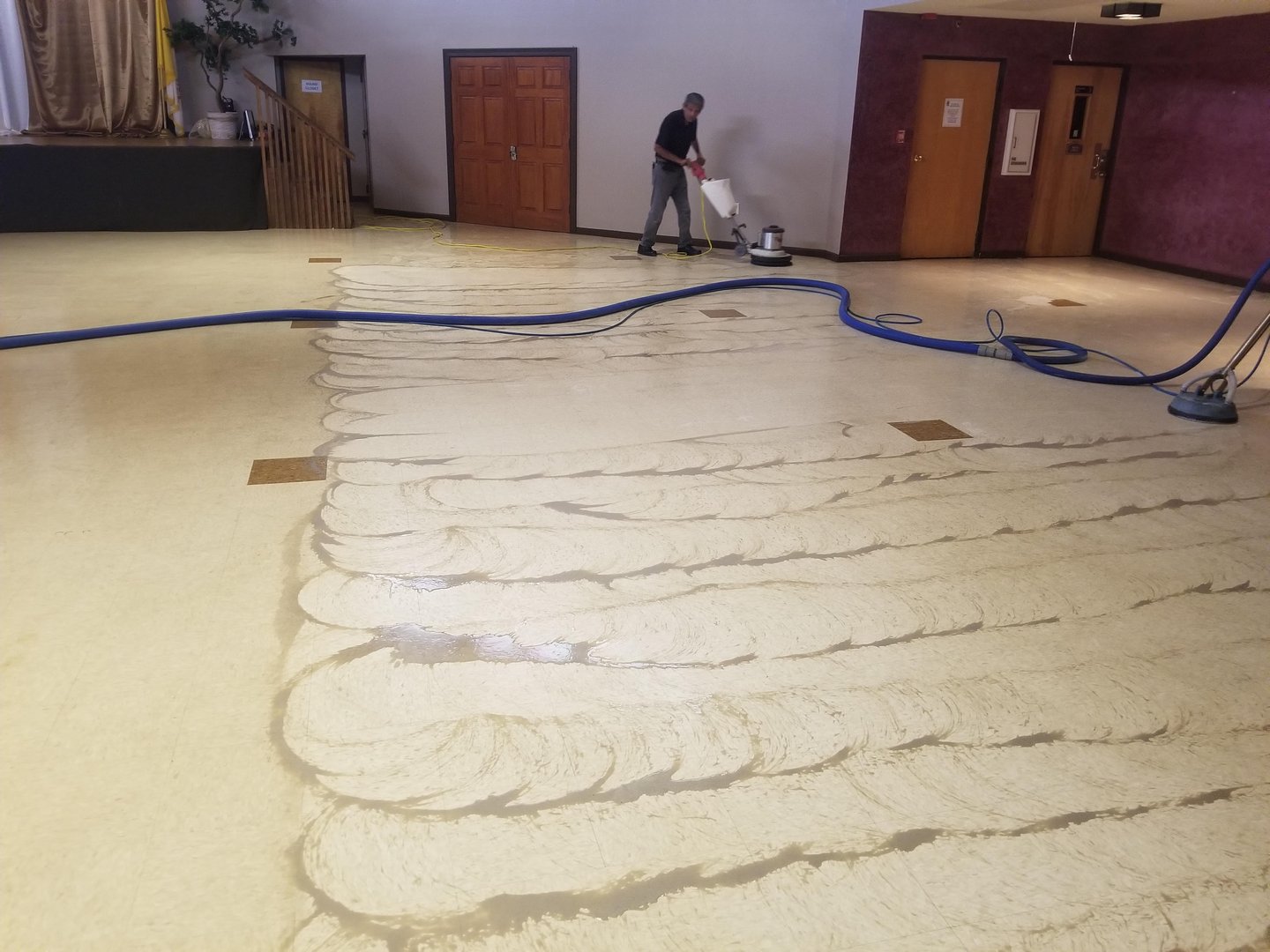 Technician stripping a large hall vinyl floor with a floor machine, swirl patterns and blue hoses visible