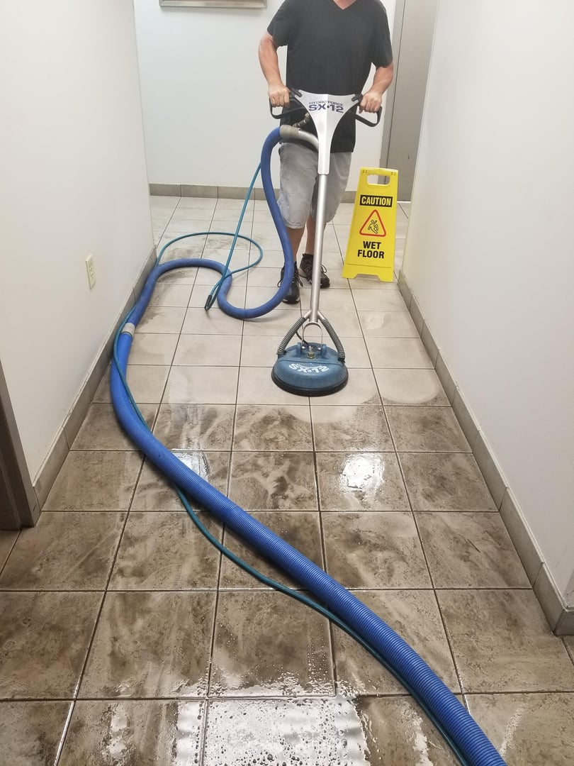 Technician using an SX-12 surface cleaner on dirty hallway tiles beside a wet floor sign and hoses