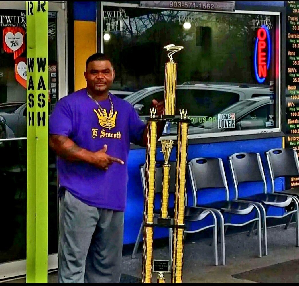The E Smooth owner in a purple E Smooth shirt holding a tall trophy outside the car wash storefront