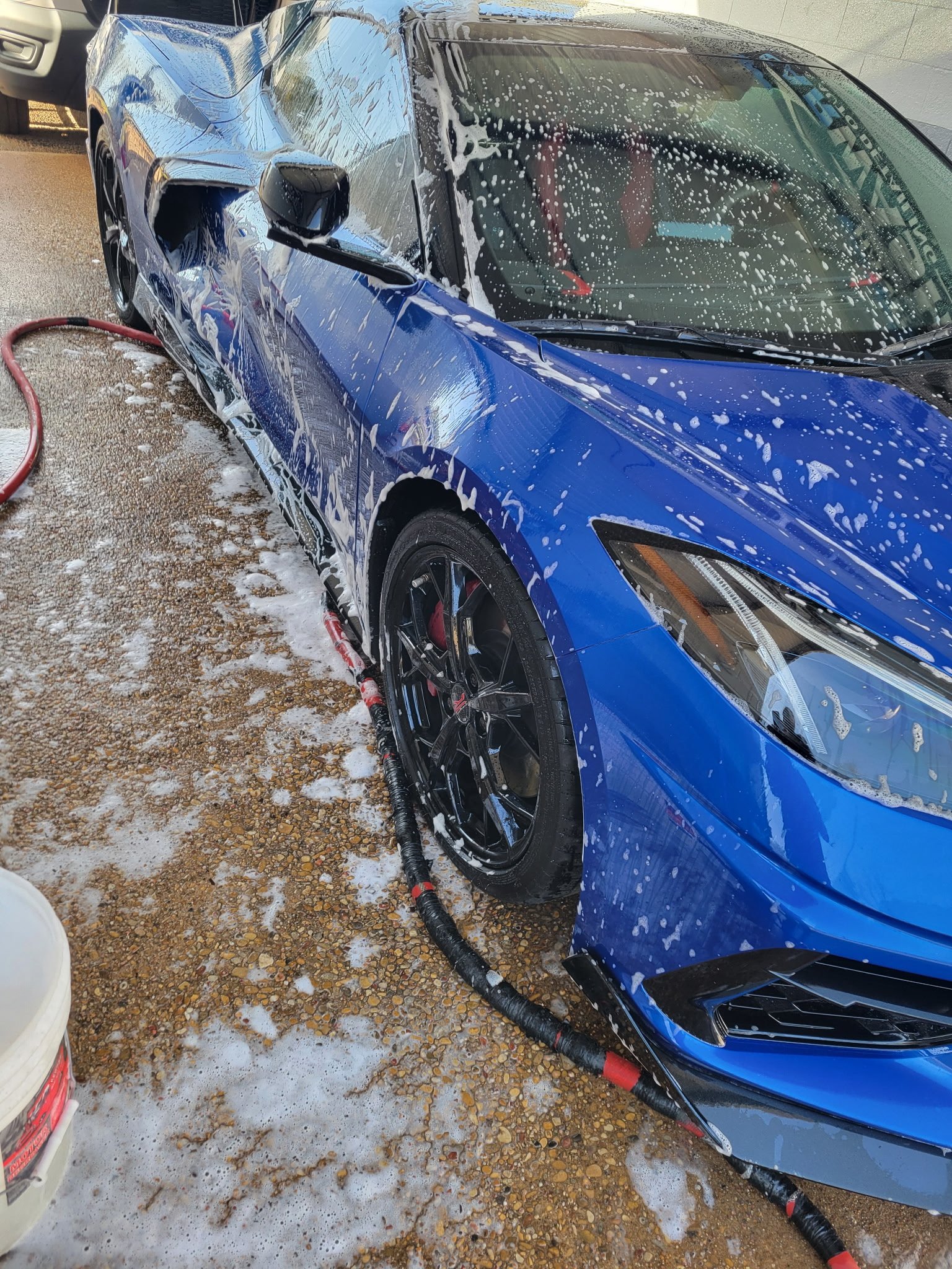 Blue Corvette covered in soap suds being hand washed with a hose and bucket on the E Smooth gravel lot