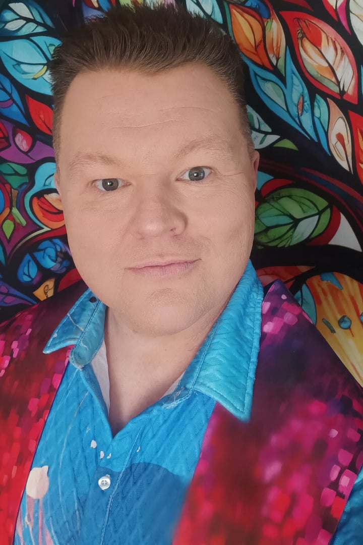Nicholas Charles smiling in a blue shirt and colorful jacket before a stained-glass tree