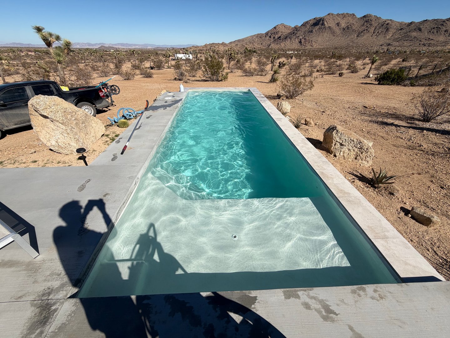 Long clear desert lap pool with a pool vacuum hose and pole on the deck and a service truck nearby