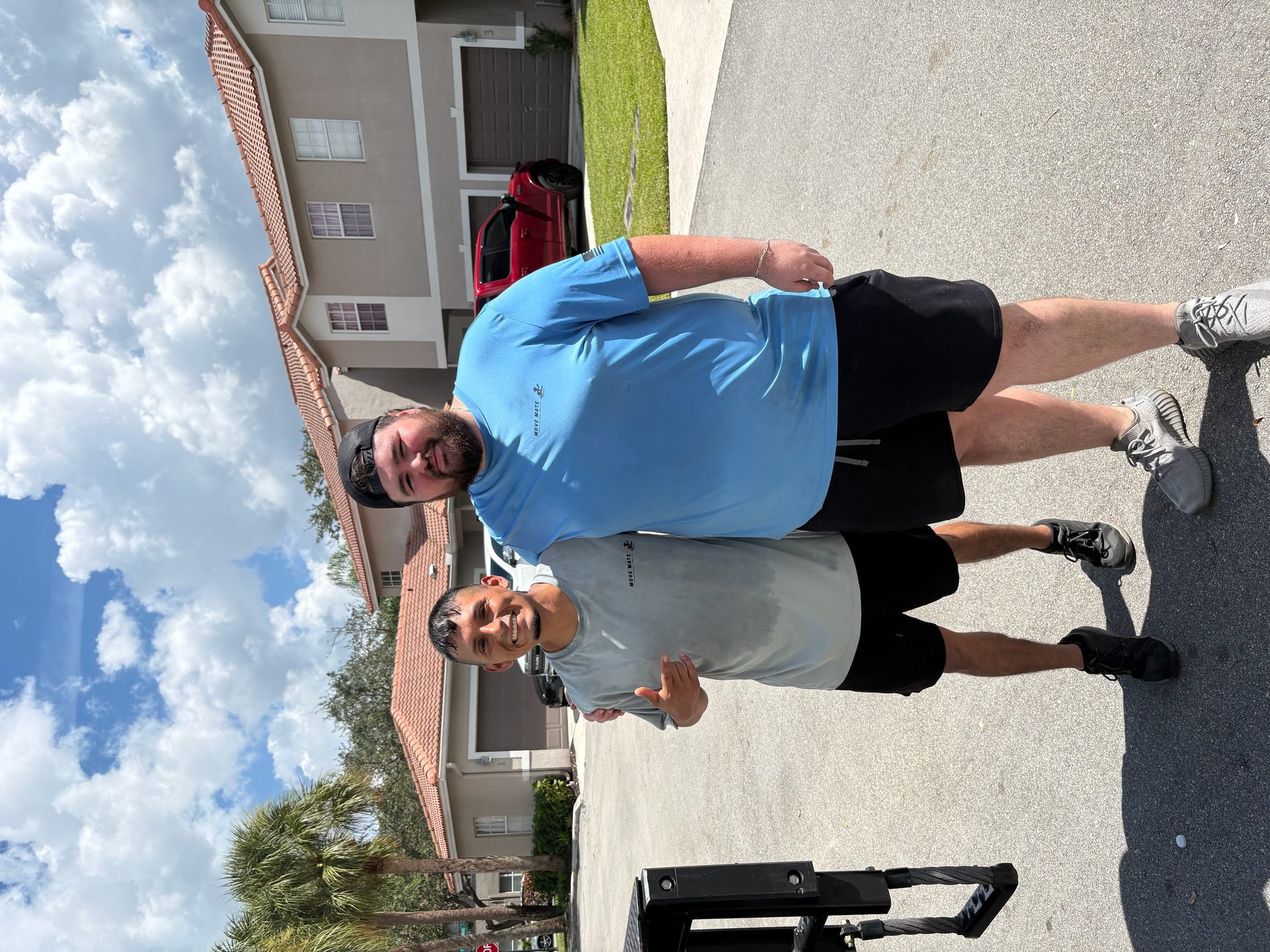 Two smiling Move Mate movers in blue and gray shirts standing on a residential driveway
