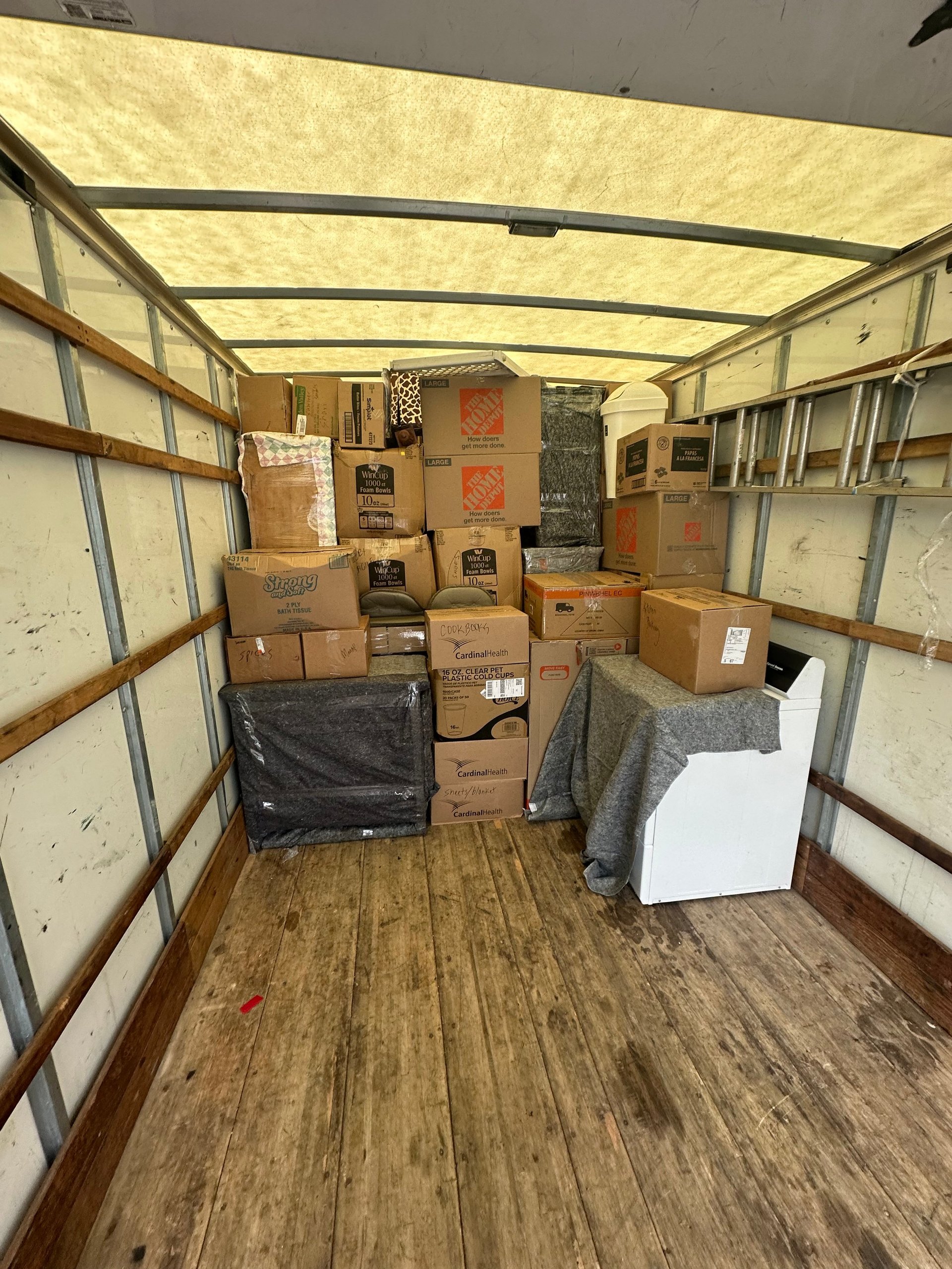 Inside a moving truck: stacked cardboard boxes, blanket-covered items and a washer on a wooden floor