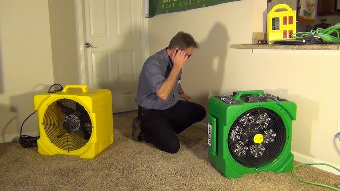 Man kneeling between a yellow GreenTech air mover and a green bed bug heater, demonstrating the heat equipment in a room
