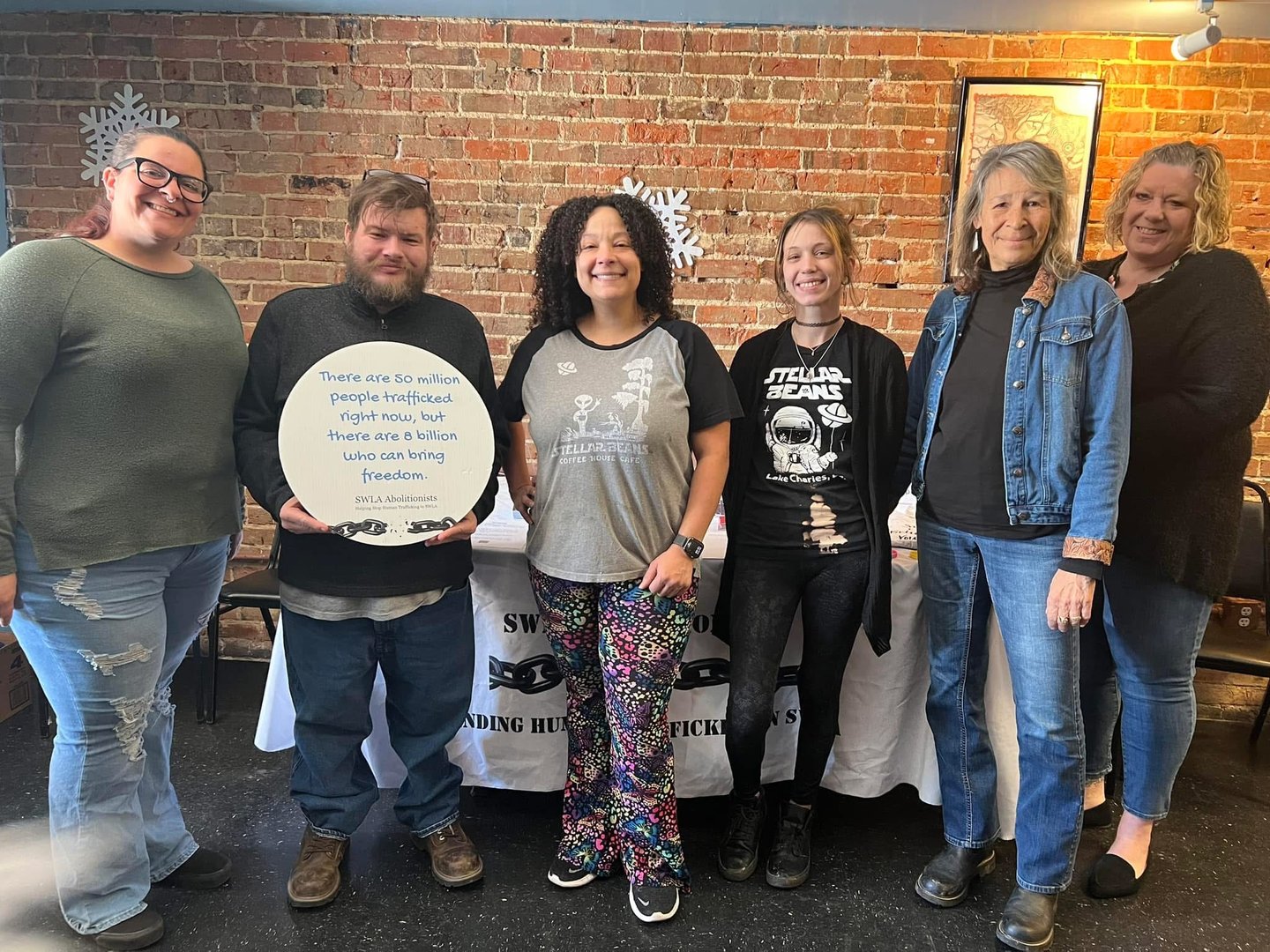 Six SWLA Abolitionists volunteers smiling together in a brick-walled coffee shop, one holding a round awareness sign about human trafficking