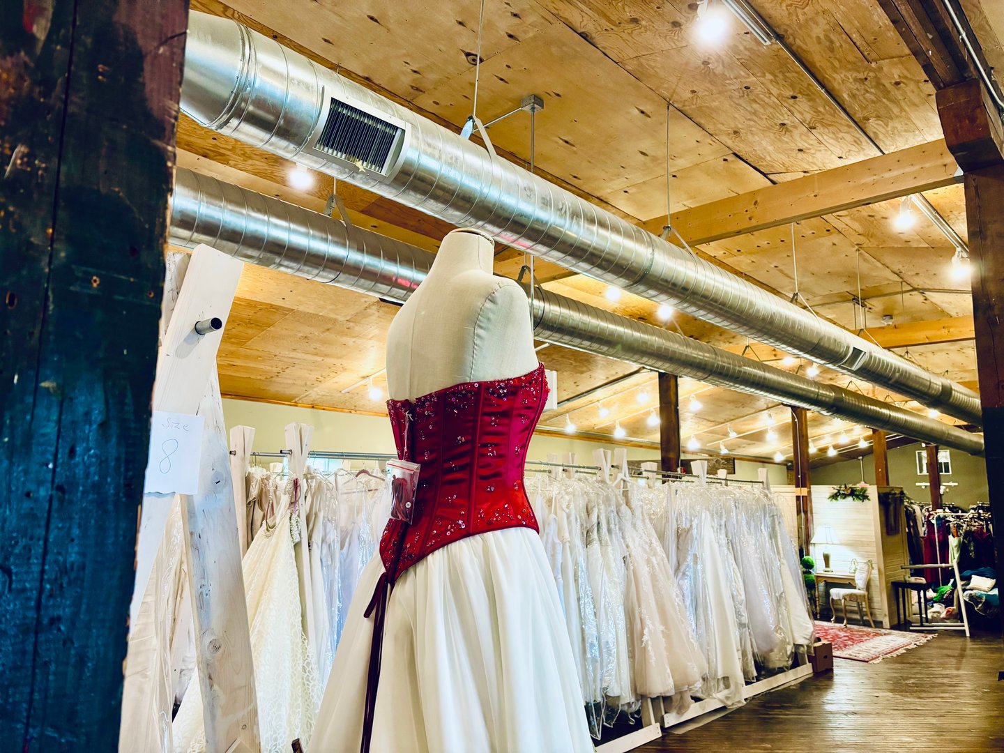 Red corset gown on a dress form in front of long racks of wedding dresses under an exposed ductwork ceiling