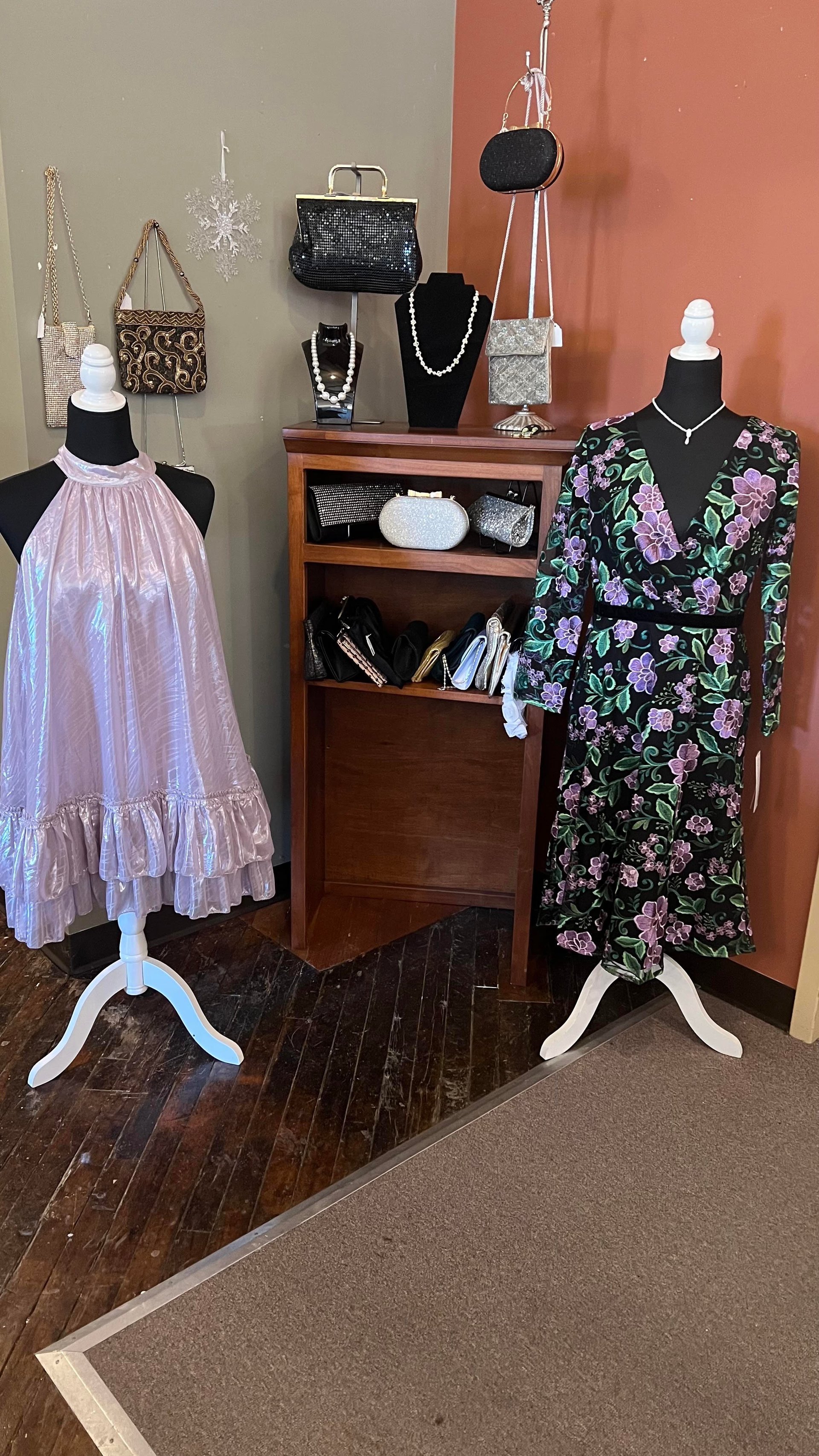 Shop corner with a lilac ruffled dress and a floral black dress on mannequins beside a shelf of handbags and jewelry