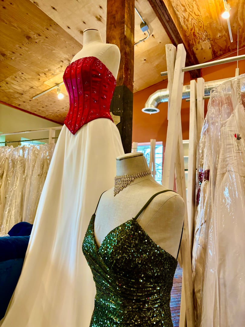 Red corset bridal gown and green sequin dress on dress forms under a wood ceiling